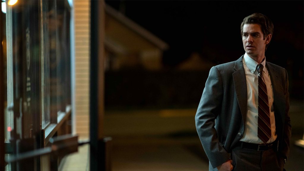 A man in a suit stands outside a building at night, looking to his right with a serious expression.