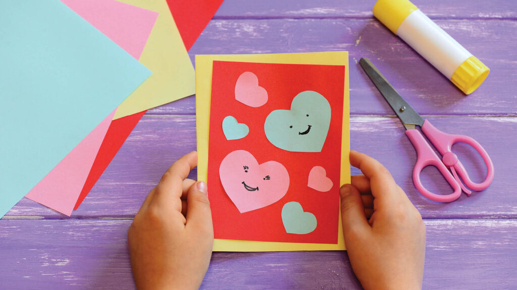 Child holds a handmade card with colorful heart cutouts, scissors, glue, and paper on a purple table.