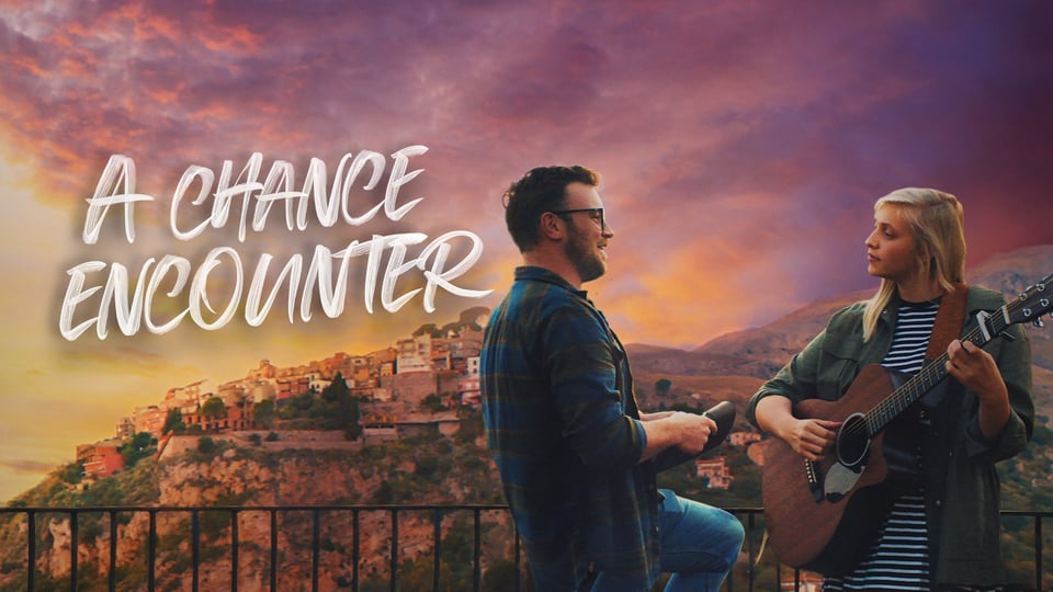 A man and woman with a guitar converse on a balcony at sunset, with a scenic village in the background. Text reads "A Chance Encounter.