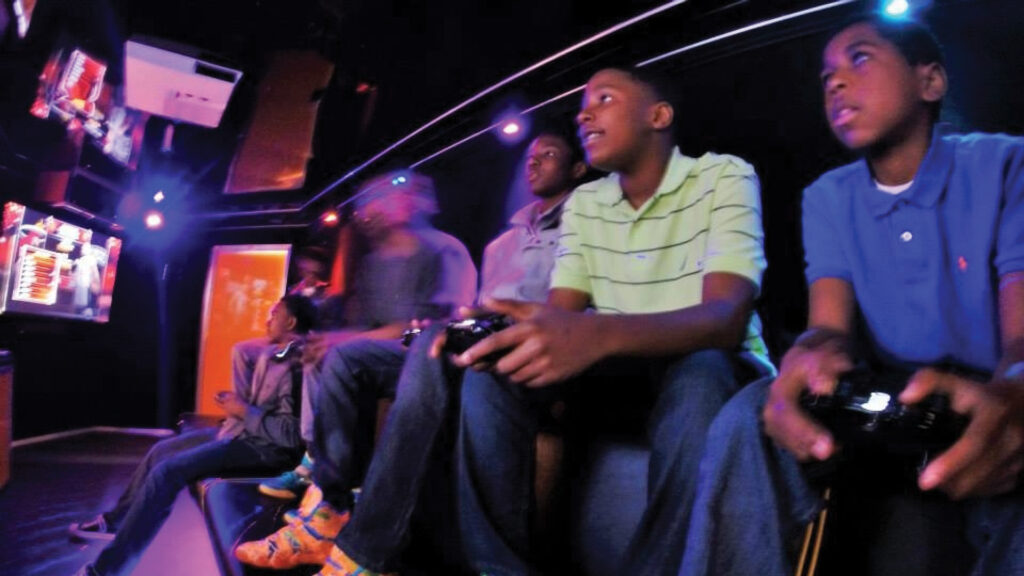 Five boys sit closely together in a dim room, playing video games with controllers, focused on screens.