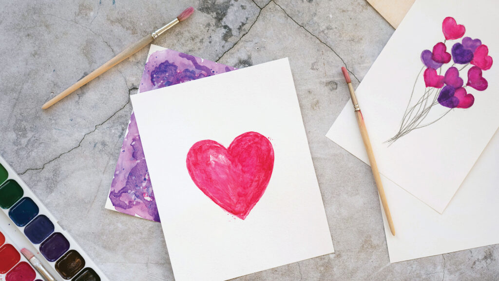 A watercolor painting of a red heart on paper, with brushes and other heart-themed art supplies nearby.