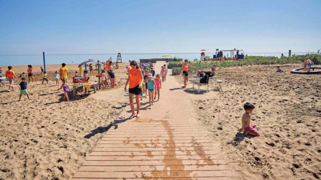Children and adults walk and play on a sandy beach with a wooden pathway under a clear blue sky.