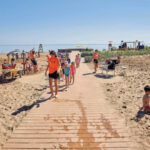 Children and adults walk and play on a sandy beach with a wooden pathway under a clear blue sky.
