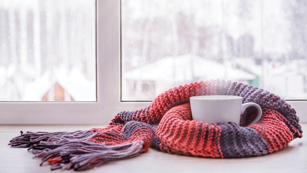 A steaming mug nestled in a cozy red and purple knitted scarf by a snowy window.