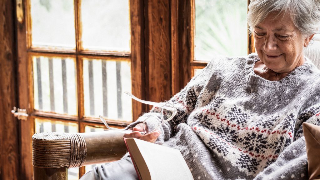 A woman with gray hair and a patterned sweater reading a book in a cozy chair by a window.