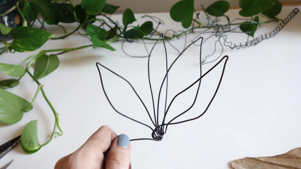 A hand holds wire shaped like leaves, with a plant vine and scissors on a white surface in the background.