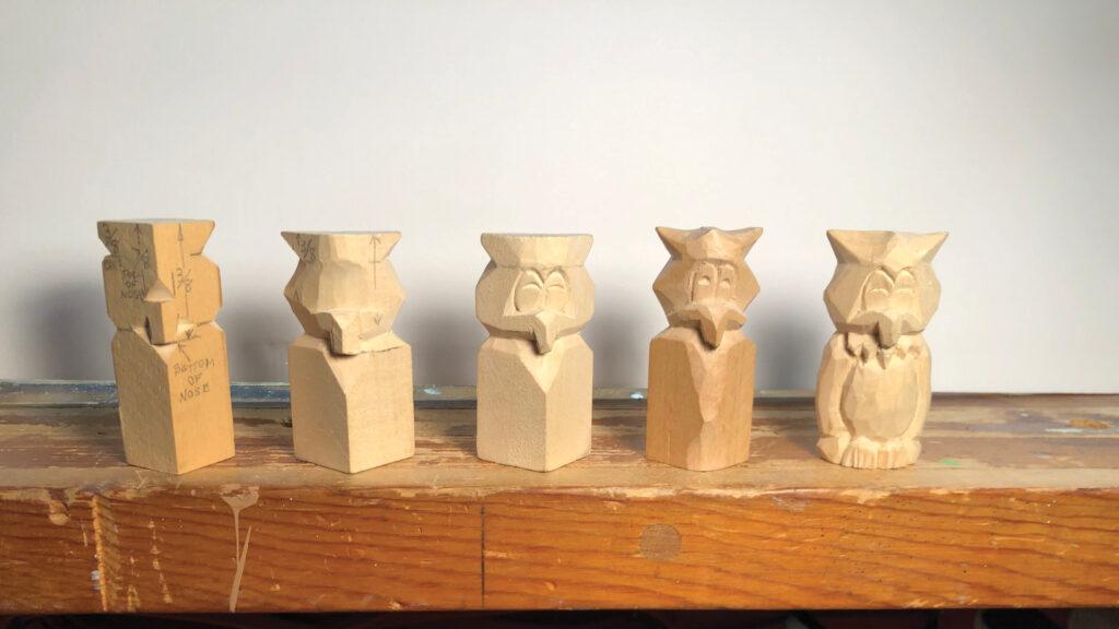 Five unfinished wooden owl carvings lined up on a wooden surface against a plain background.