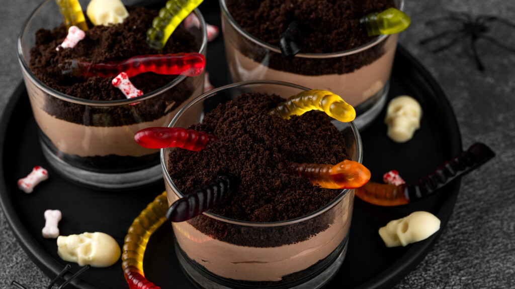 Chocolate mousse cups topped with crushed cookies, gummy worms, and candy bones for a spooky Halloween treat.