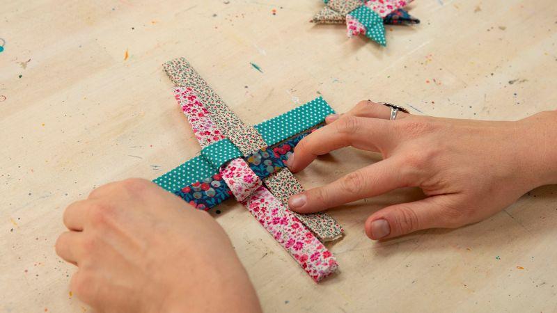 Hands weaving colorful fabric strips into a patterned decoration on a wooden table.