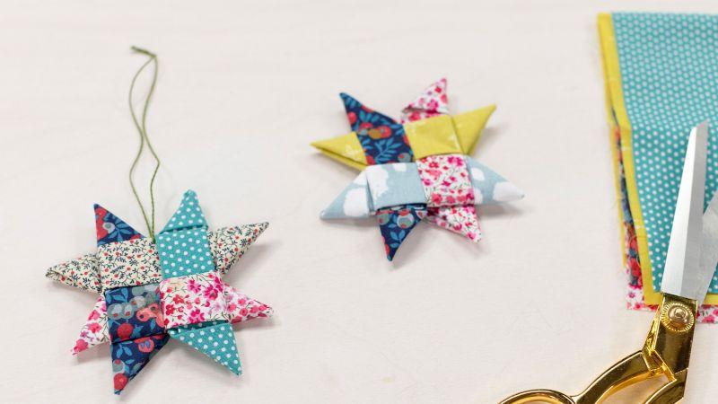 Two colorful folded fabric stars on a white surface, next to gold scissors and fabric pieces.