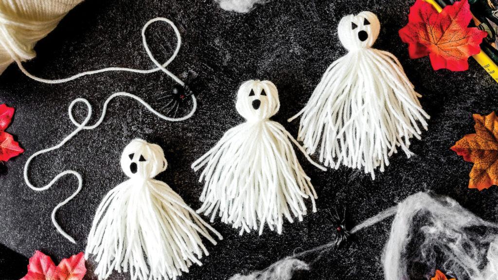 Three yarn ghosts with black eyes and mouths on a dark surface, surrounded by fake cobwebs and autumn leaves.