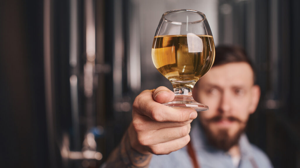 A person holding a glass of golden beer toward the camera, with a blurred background.